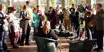 Woman in chair with back to camera talks to journalists with notepads, cameras.
