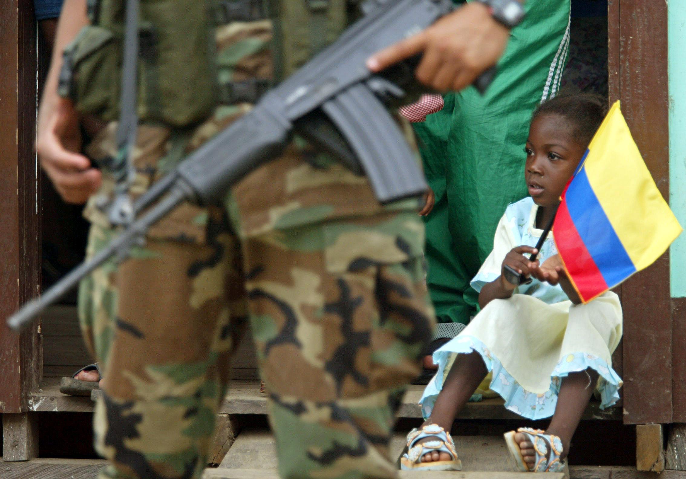 Una niña senrada en el suelo con una bandera de Colombia. Ante ella, vista parcial de un soldado armado.