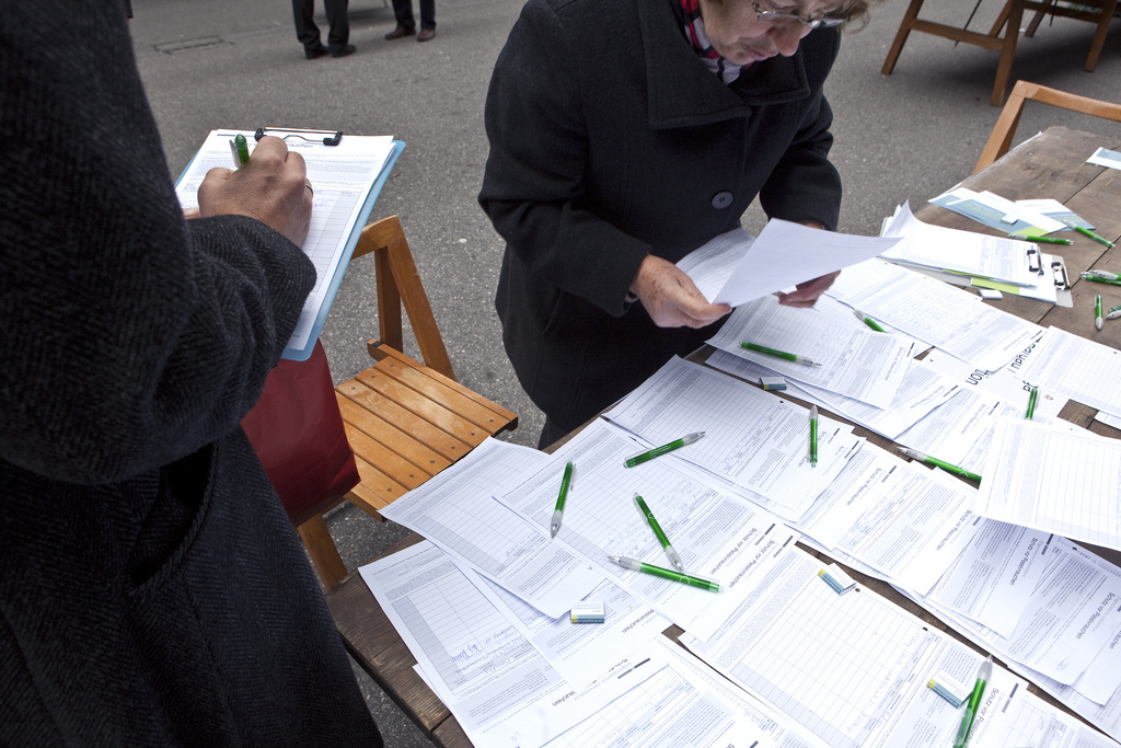 Una mesa con papeles y plumas, una persona leyendo un documento y otra firmando