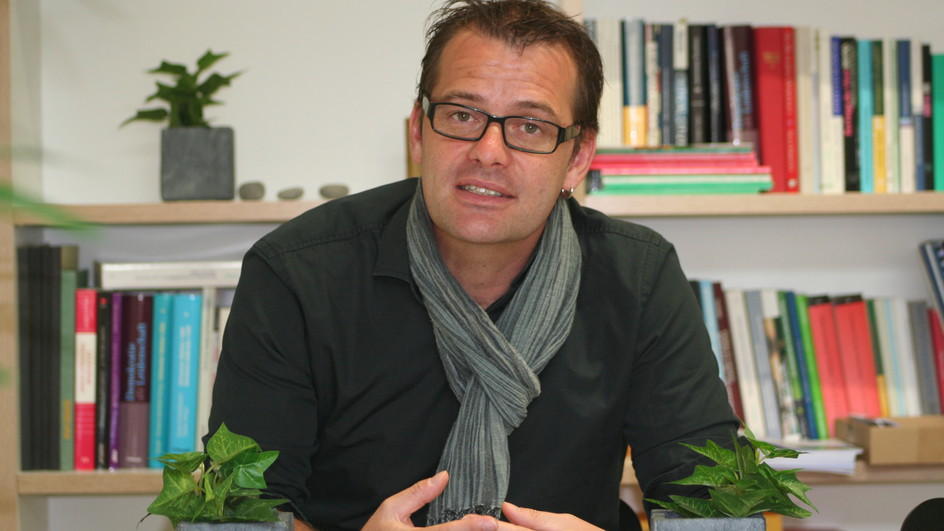 Marc Bühlmann sitzt im schwarzen Pullover und mit grauem Schal an seinem Schreibtisch