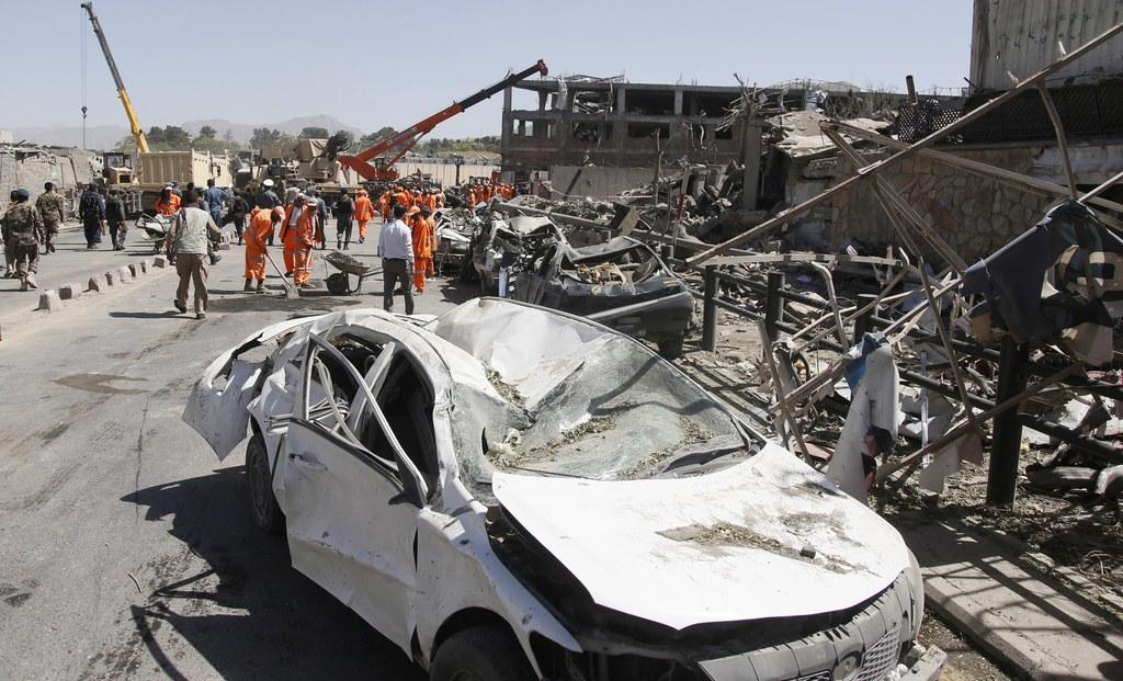 A twisted white car stands next to the ruins of a building, with cranes and workers clearing away debris in the background