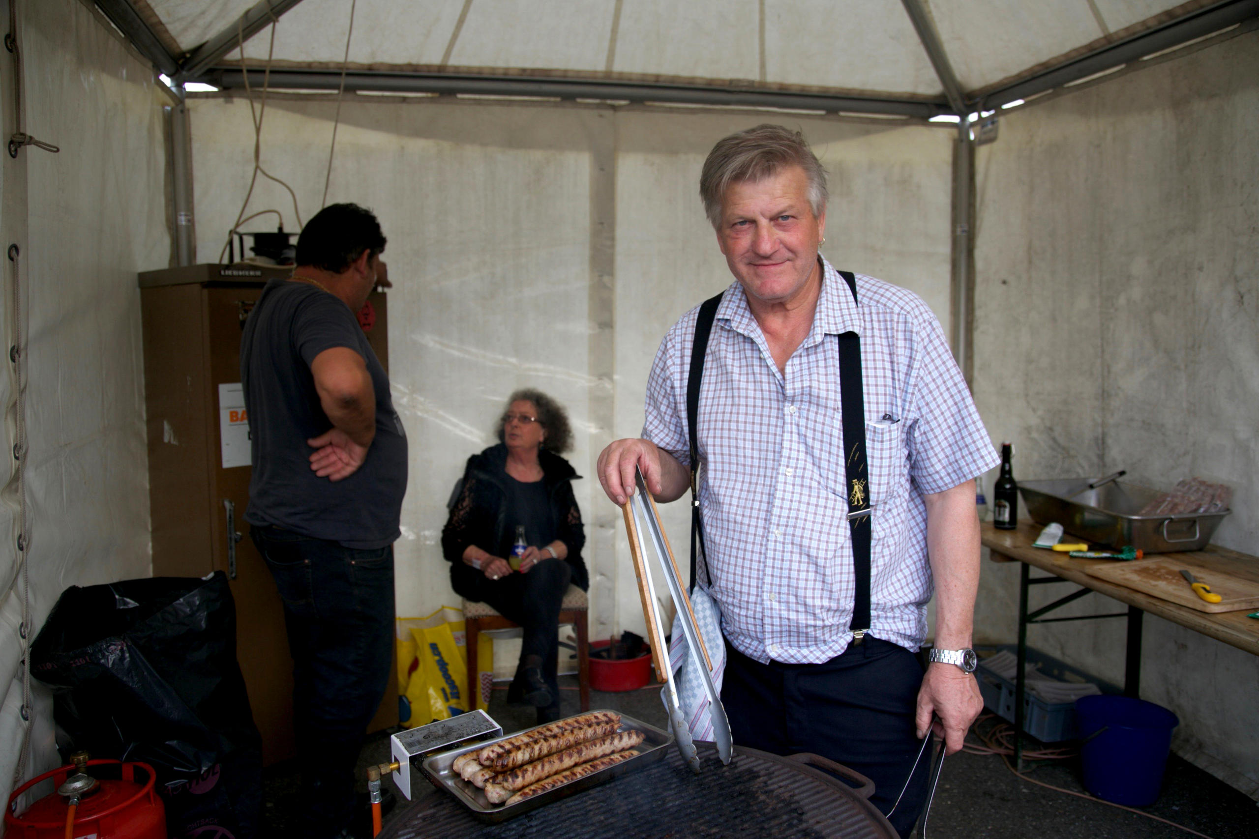 A man stands stands next to a grill holding grill tongs, two people are in the background.