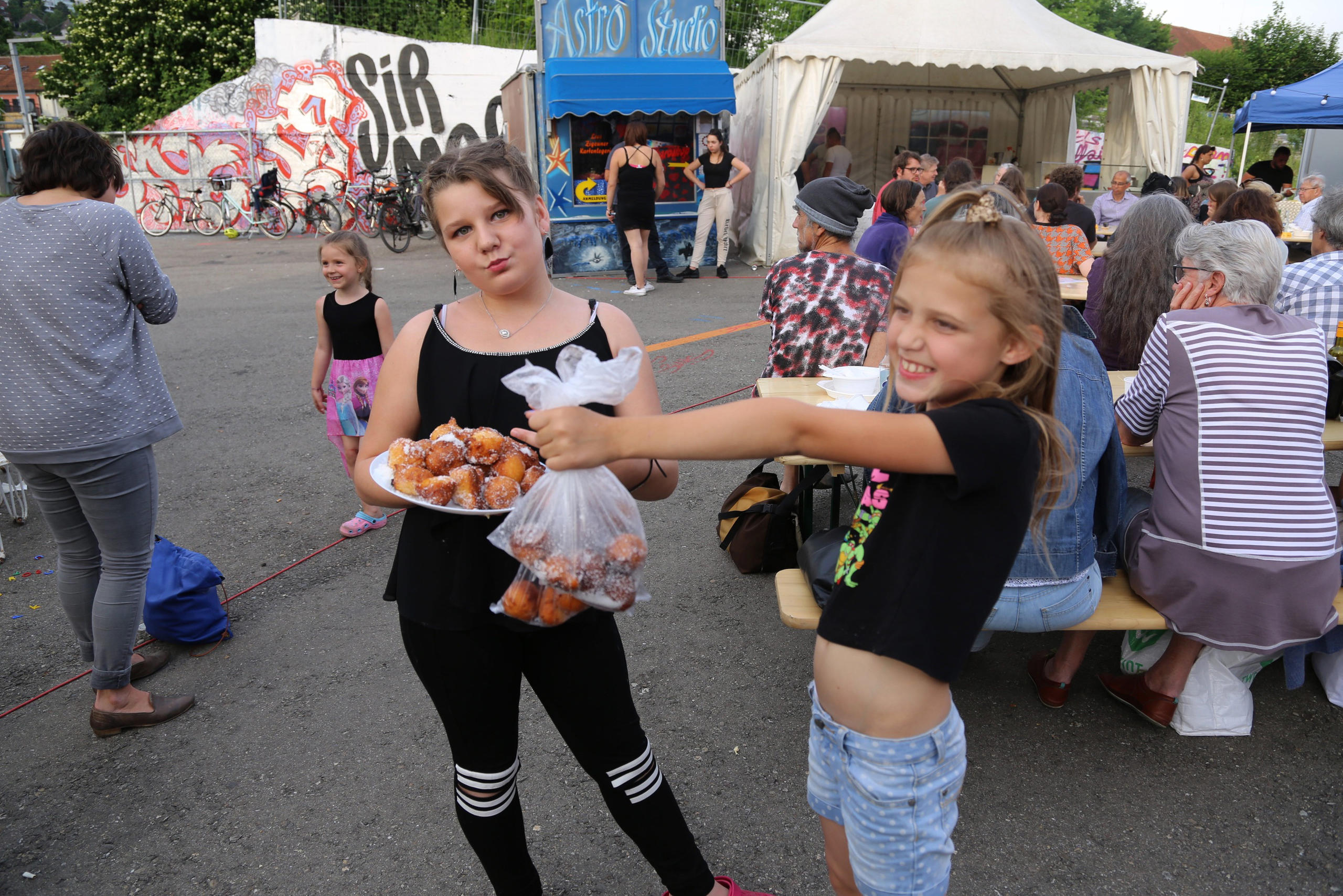 two young girls, one holding a plate of doughnuts, the other holding a bag of doughnuts. People sitting in the background