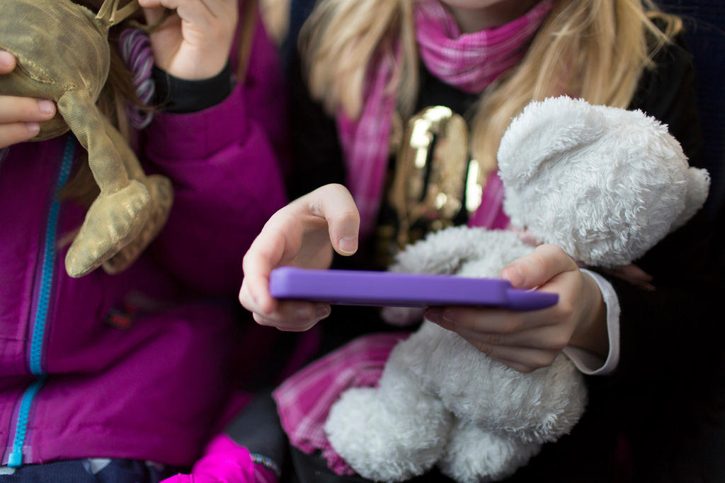 A young girl with a stuffed bear plays on her smartphone.