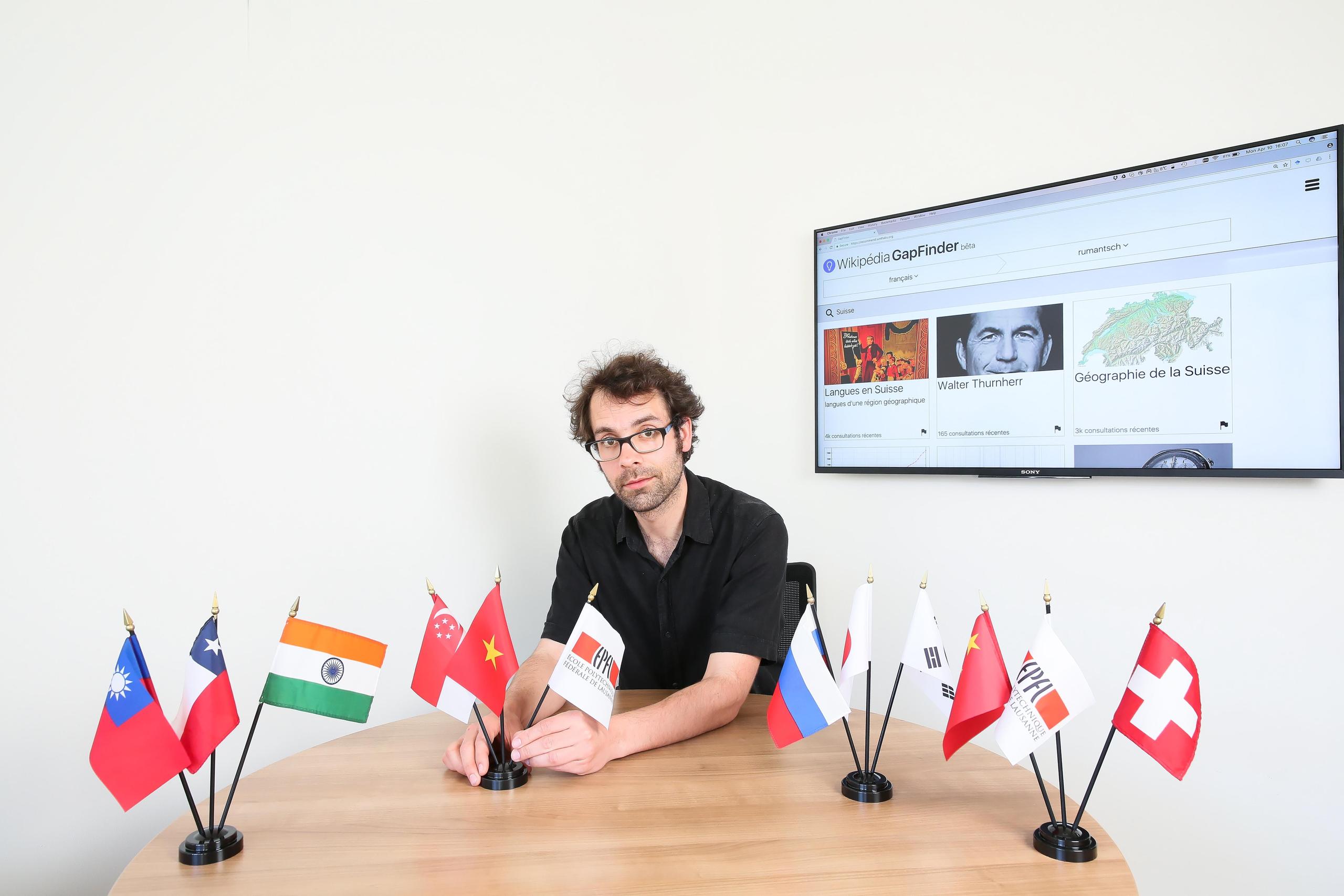 EPFL researcher Robert West poses with national flags in front of a screen showing the home page of Wikipedia GapFinder platform
