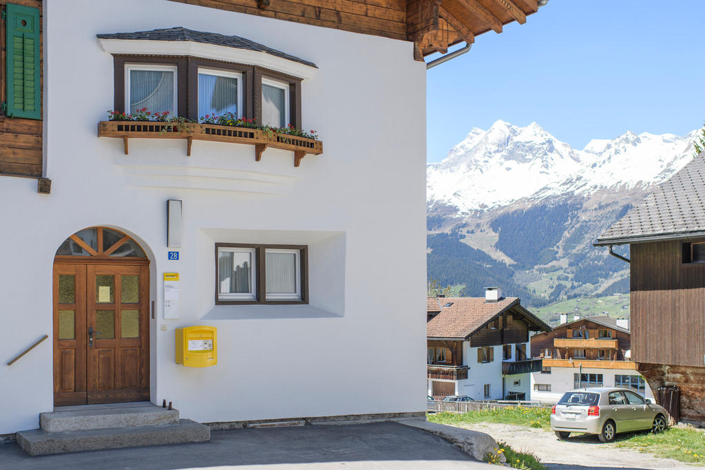 Eine Poststelle mit gelbem Briefkasten an der weissen Aussenmauer in einem Dorf, im Hintergrund sind Berge zu sehen.