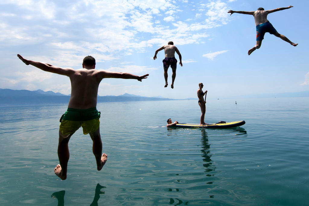 Swimmers leap in the air above Lake Geneva on a sunny day while a paddle boarder goes by