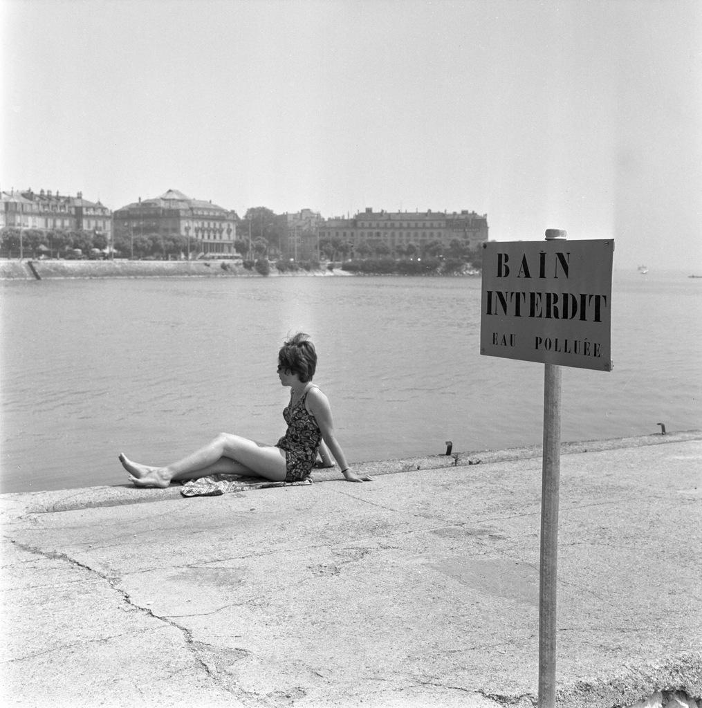 Lago de Neuchâtel em 1964: uma mulher sentada às margens ao lado de uma placa com os dizeres  Banho proibído.