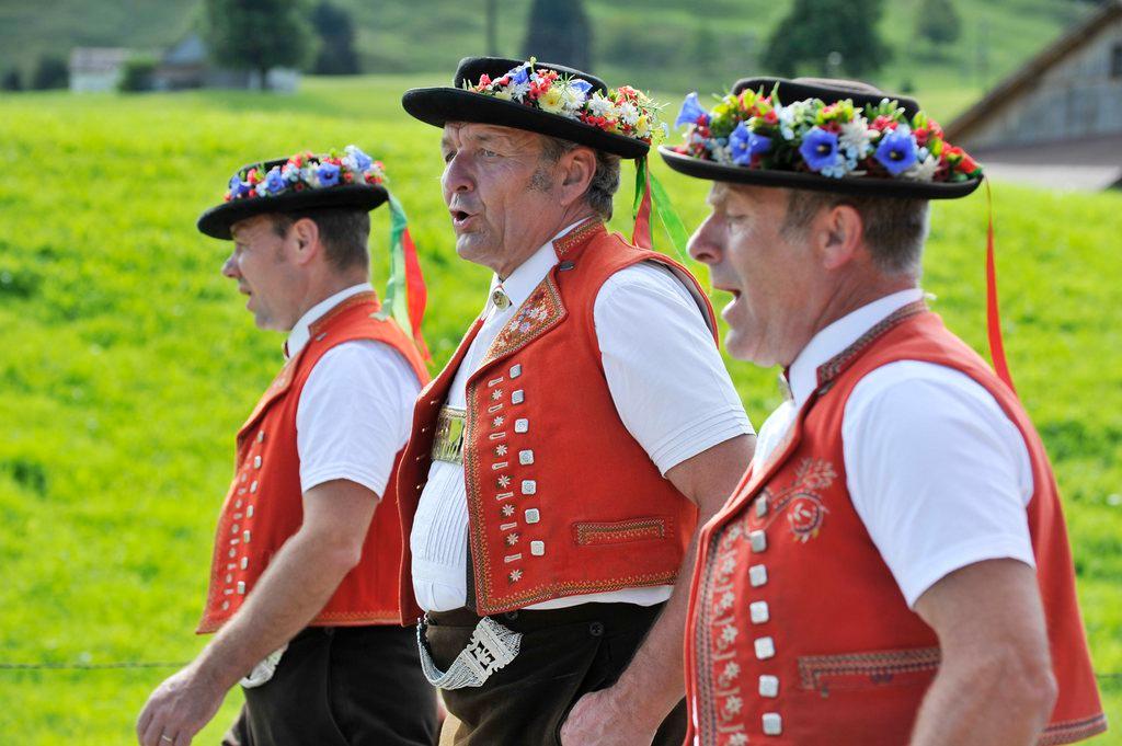 Three men in traditional Swiss dress yodel as they walk across a meadow.