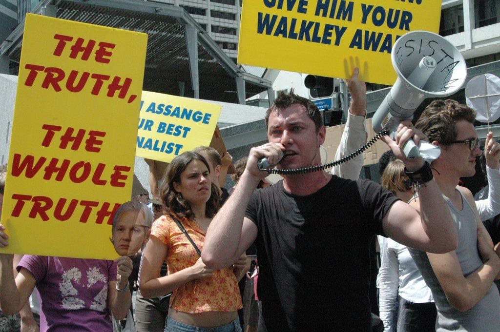 A man speaks into a megaphone amidst a crowd holding banners demanding truth