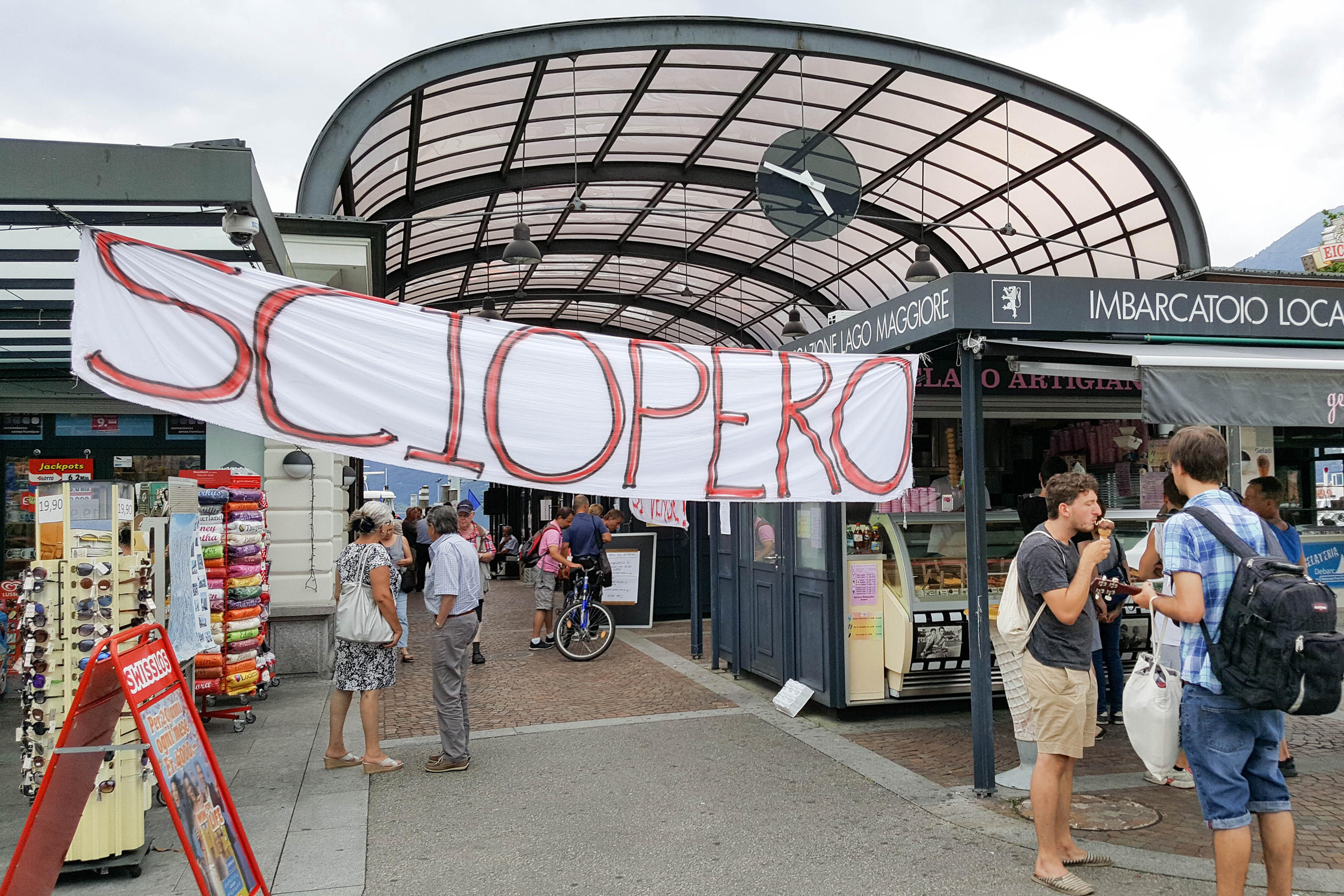 Streik steht auf einem Plakat an einer Schiffstation, Touristen essen Eis und stehen neben einem Kiosk.