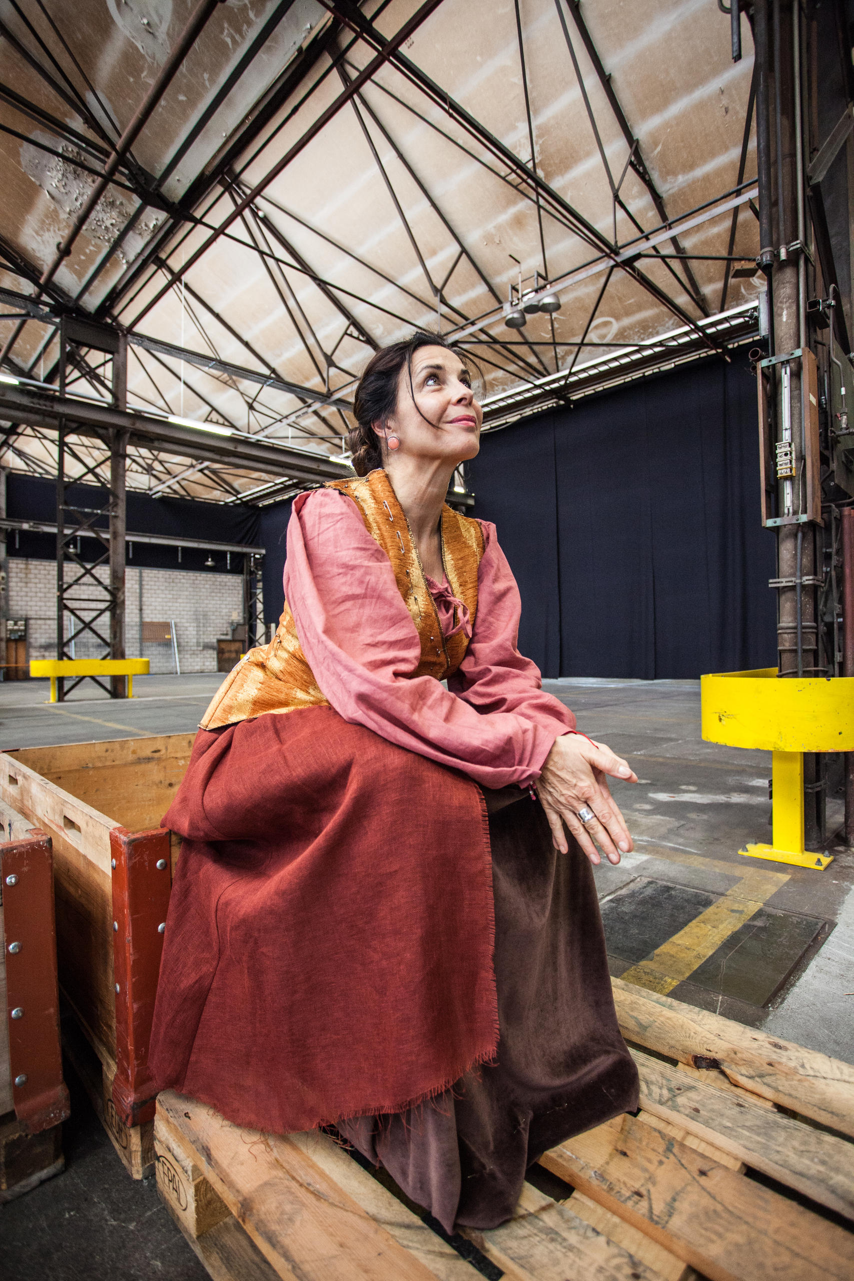 Masha Karell, wearing a red robe and gold waistcoat, sits on a wooden box on the stage.