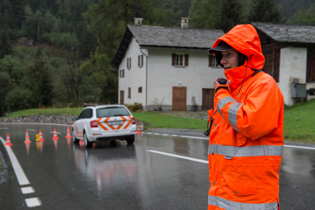 A police officer, standing in a road in wet weather next to a police car and cones, wears an orange rain and talks into jacket