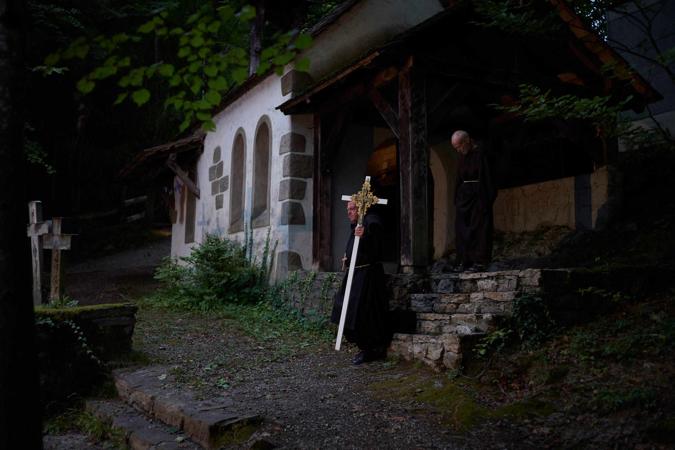 Two priests, one carrying a large white cross, walk down steps in front of a house.