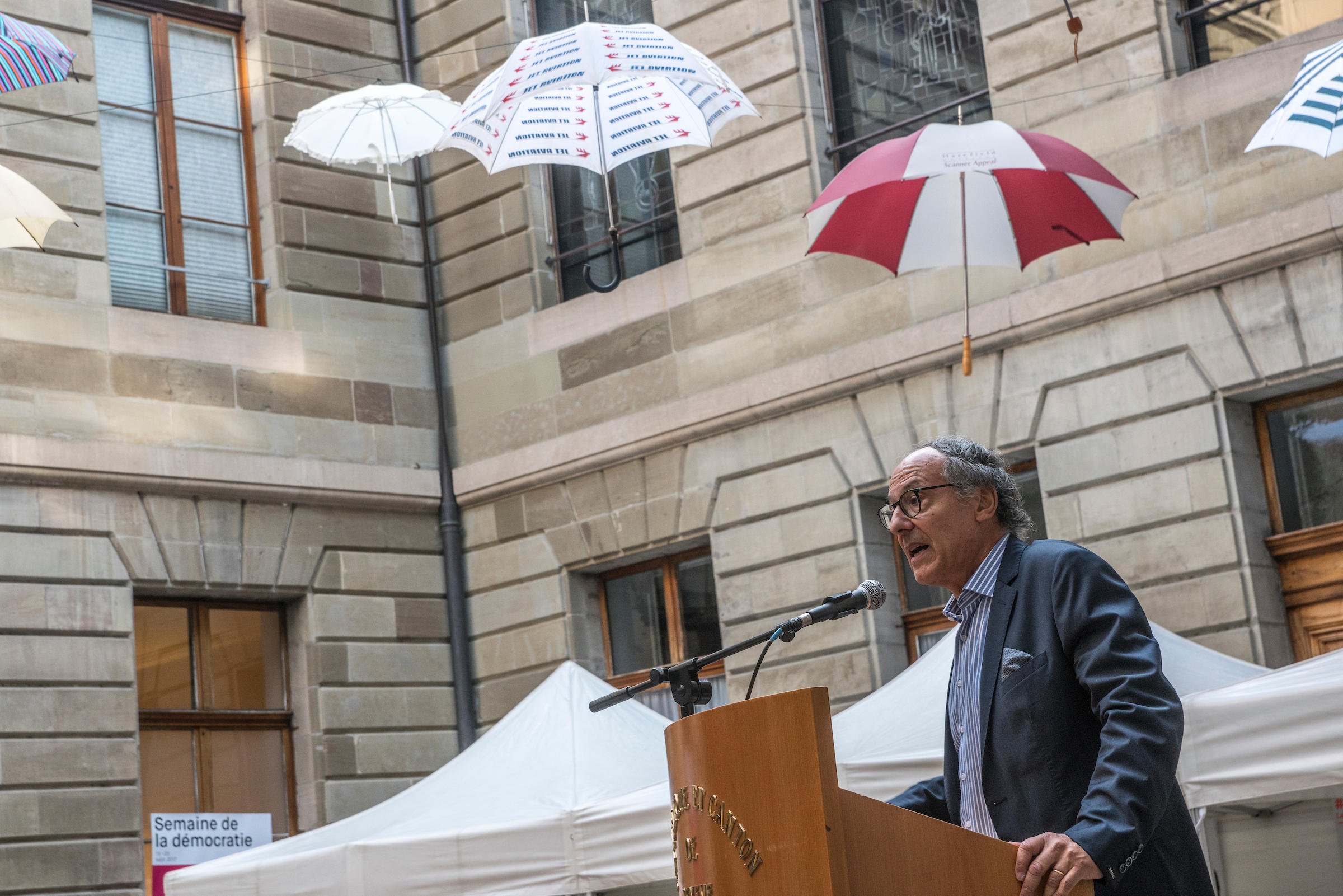 Ein Mann steht vor einem Rednerpult. Im Hintergrund sind Regenschirme künstlerisch aufgehängt.