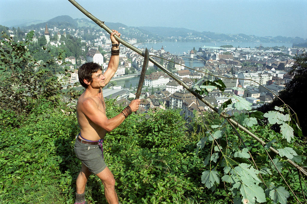 A bare chested Bruno Manser cuts a branch with a machete in the jungle with a town in the background