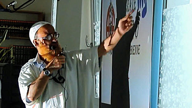 A man in white clothing, with an orange beard, speaks into a microphone whilst pointing at a wall poster