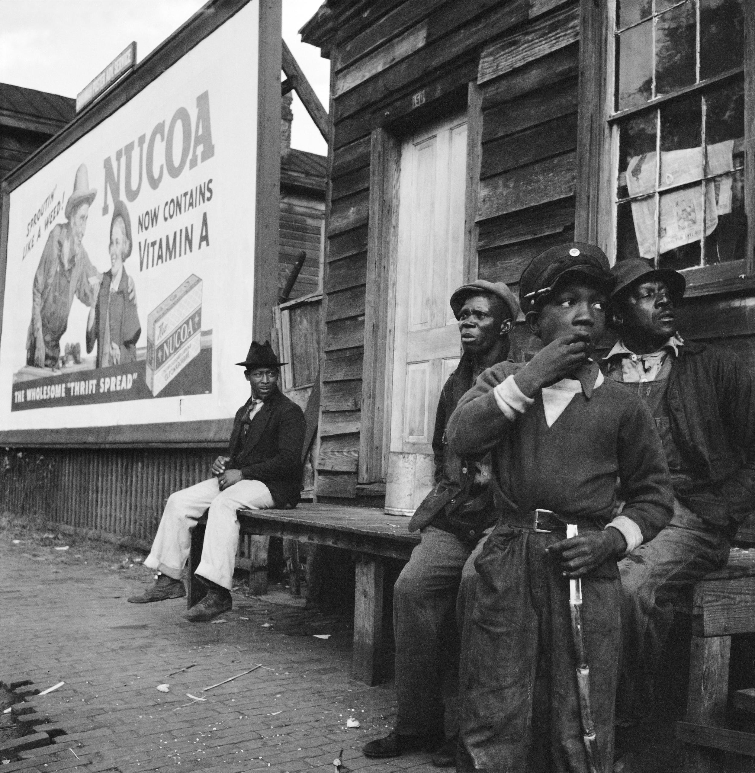 Groupe de noirs assis devant une maison en bois.