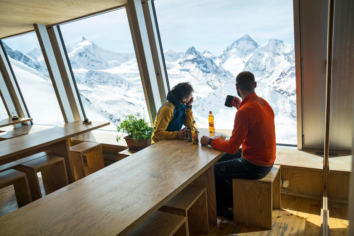 Two people sit at table looking out the window at snow-covered alpine peaks