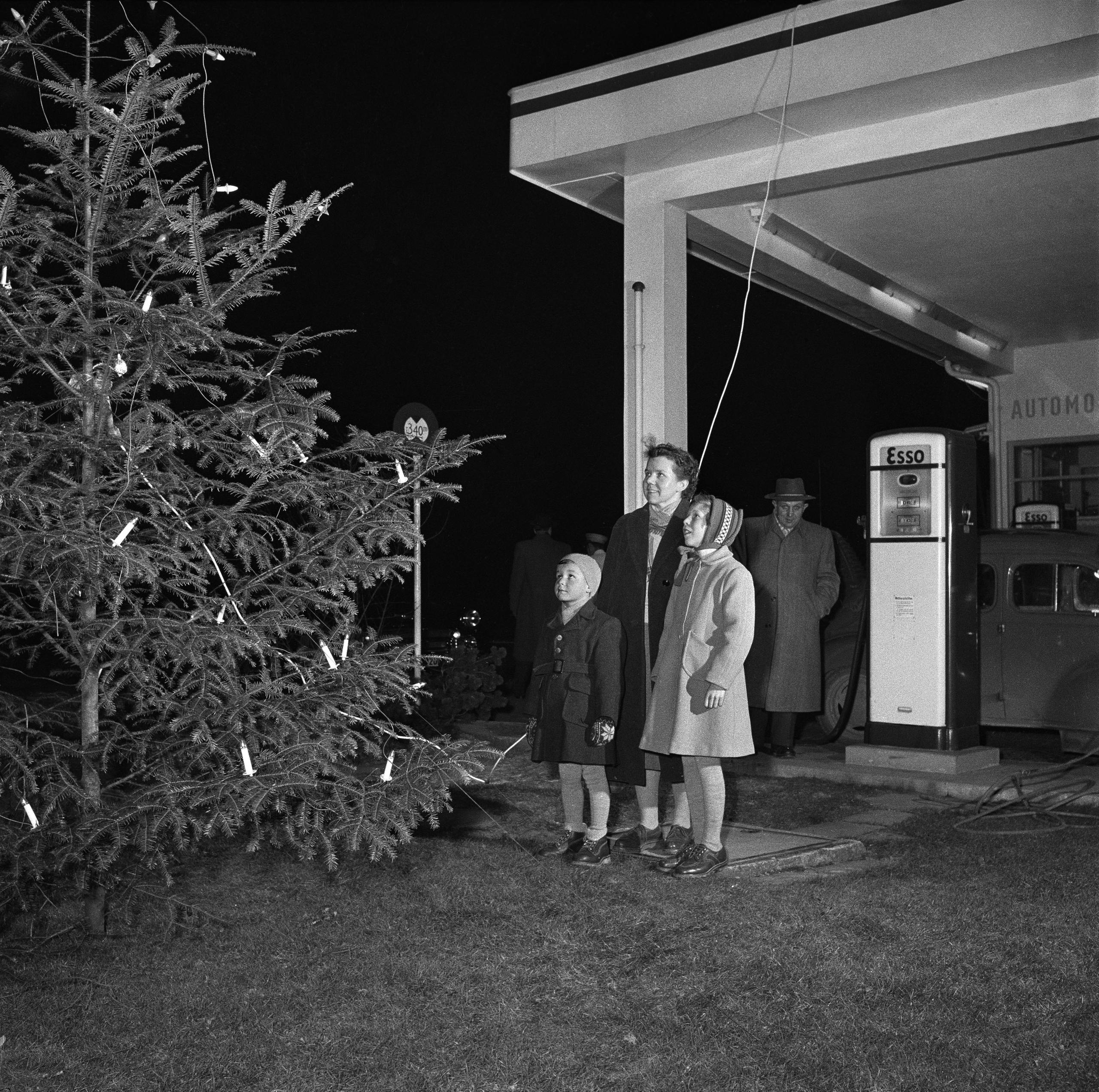 three people standing in front of a christmas tree at a petrol station