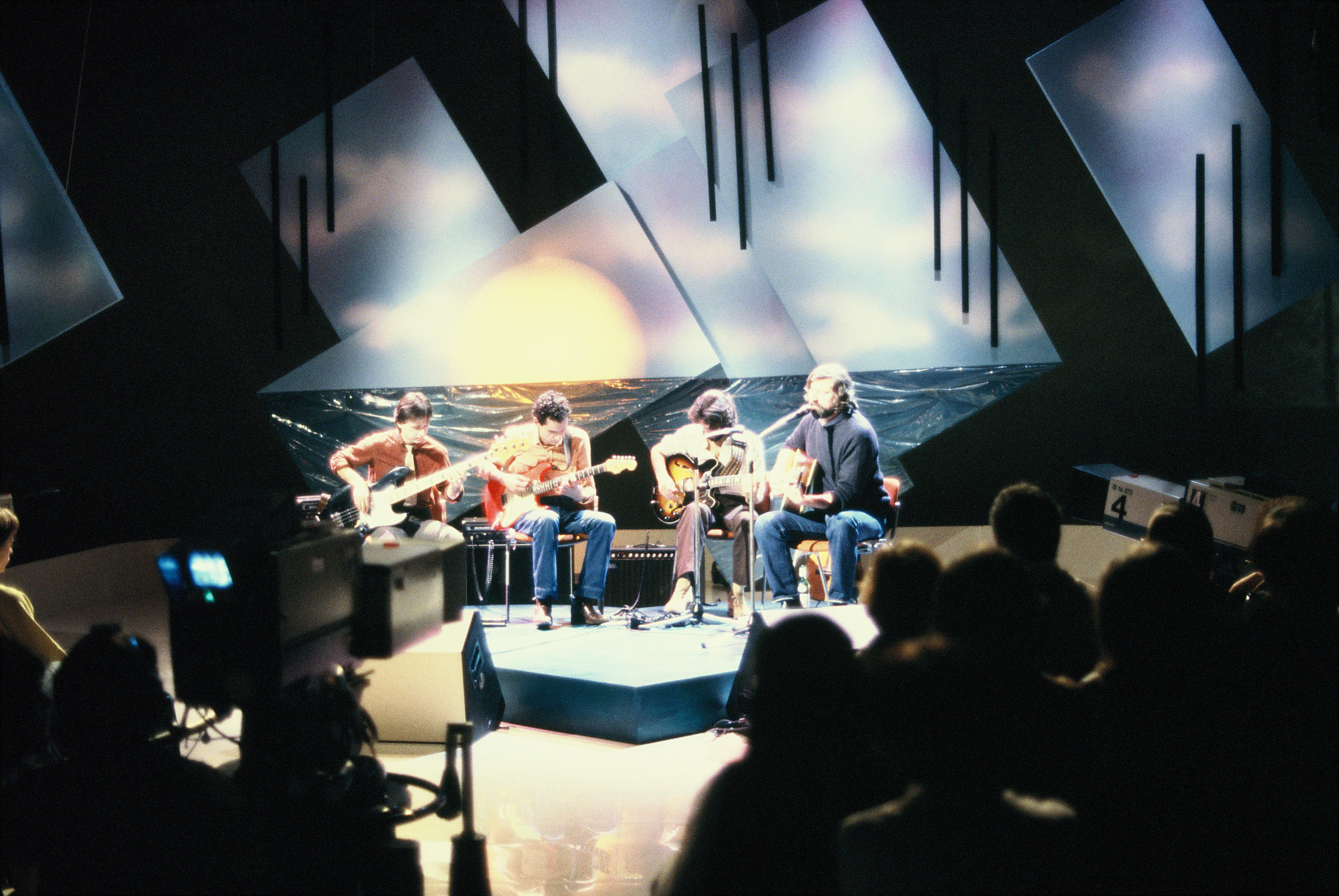 Immagini di Francesco Guccini e tre musicisti sul palco. Sullo sfondo, immagine di un tramonto.