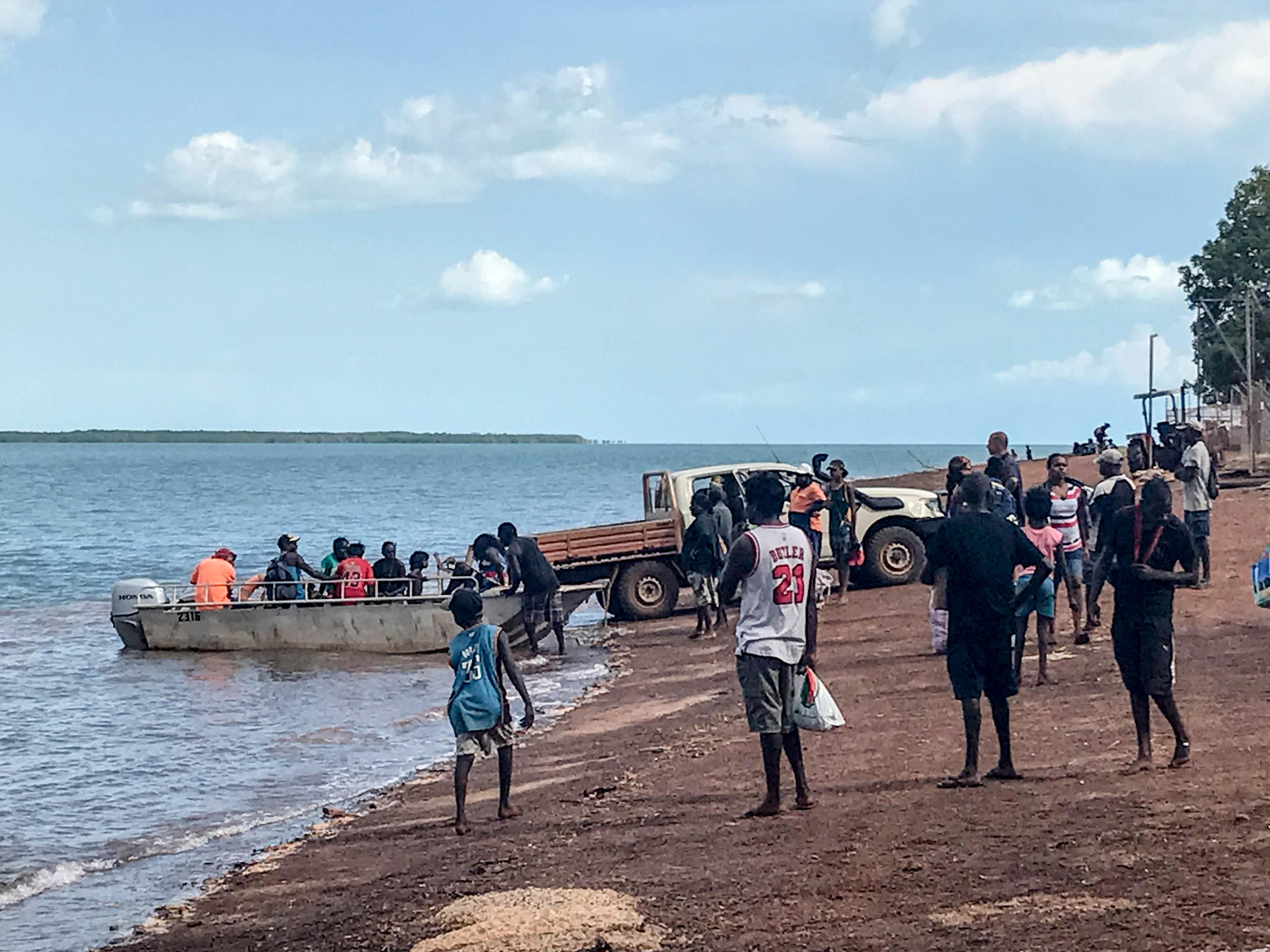 People standing on a beach with people boarding a small boat and a flat bed truck