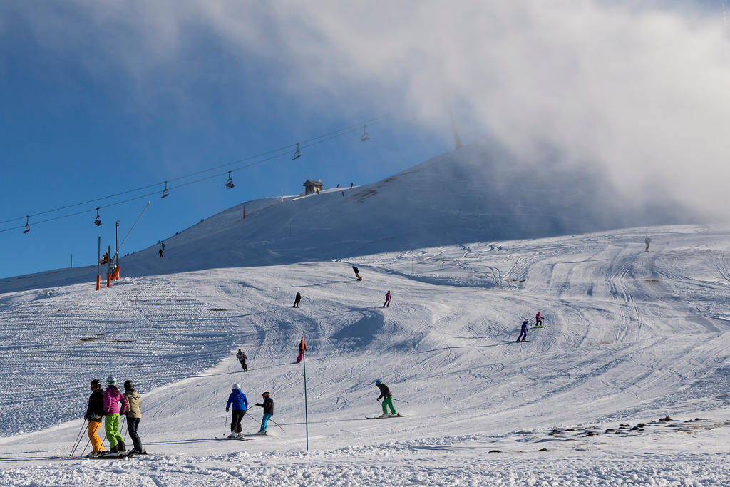 Esquiadores em uma ampla pista, com teleférico ao fundo