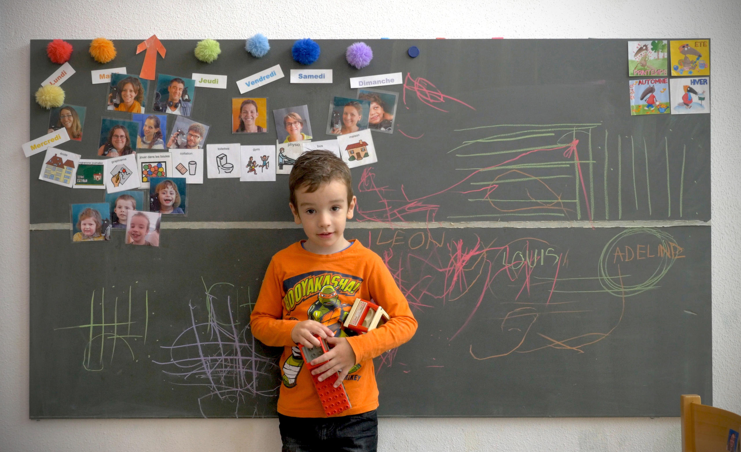 Bambino davanti a una lavagna, sulla quale sono appesi dei ritratti fotografici e disegnati alcuni scarabocchi a colori