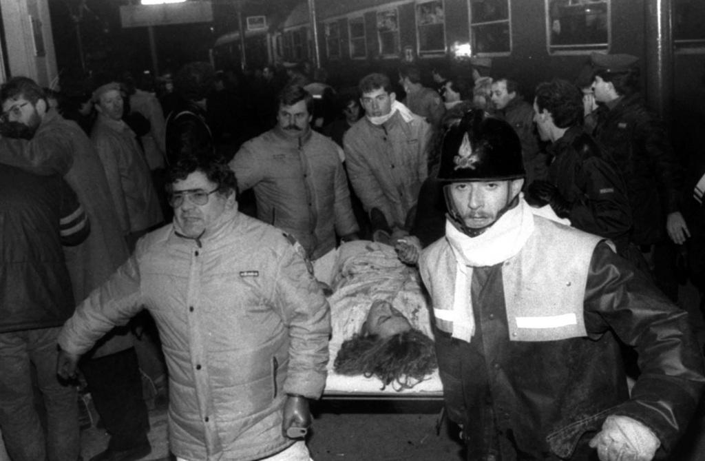 Due soccorritori trasportano una barella con una donna ferita. Sullo sfondo, treno fermo in stazione