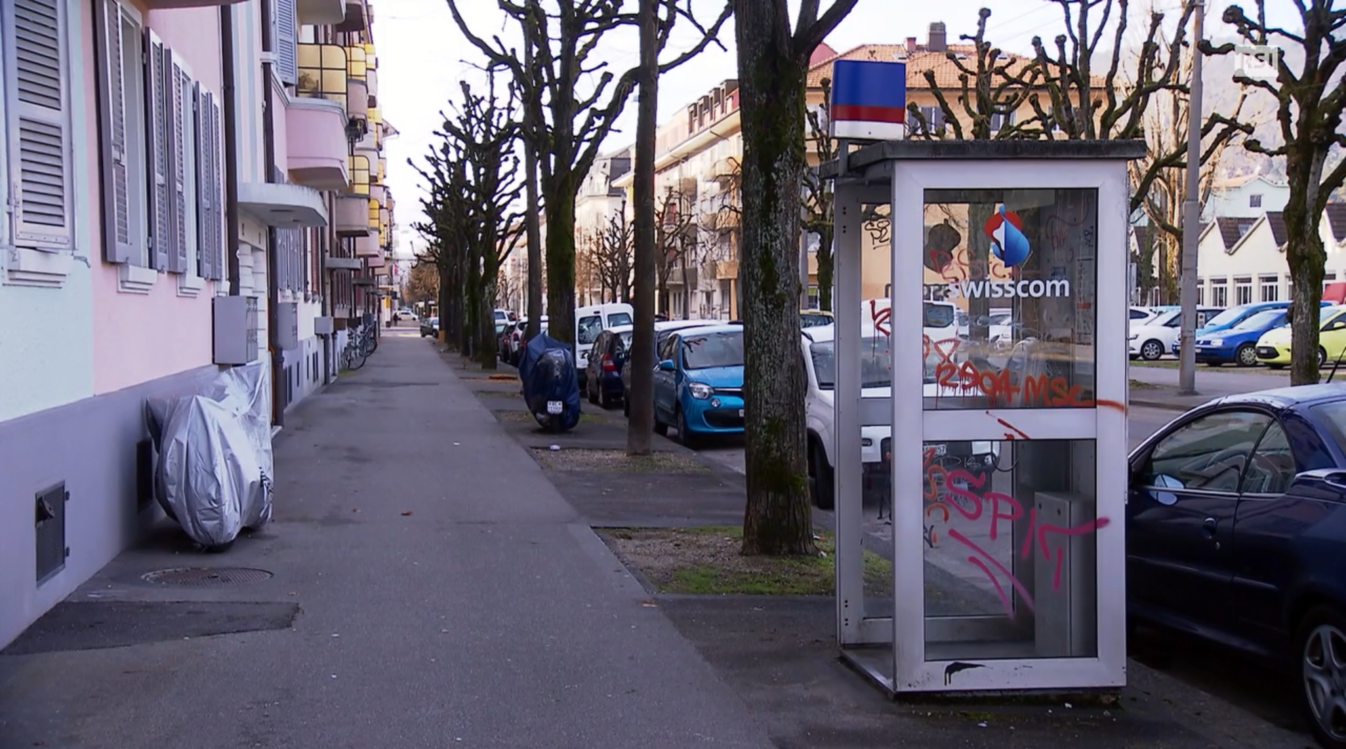 Un viale alberato con palazzi popolari, auto parcheggiate e una cabina telefonica in primo piano.