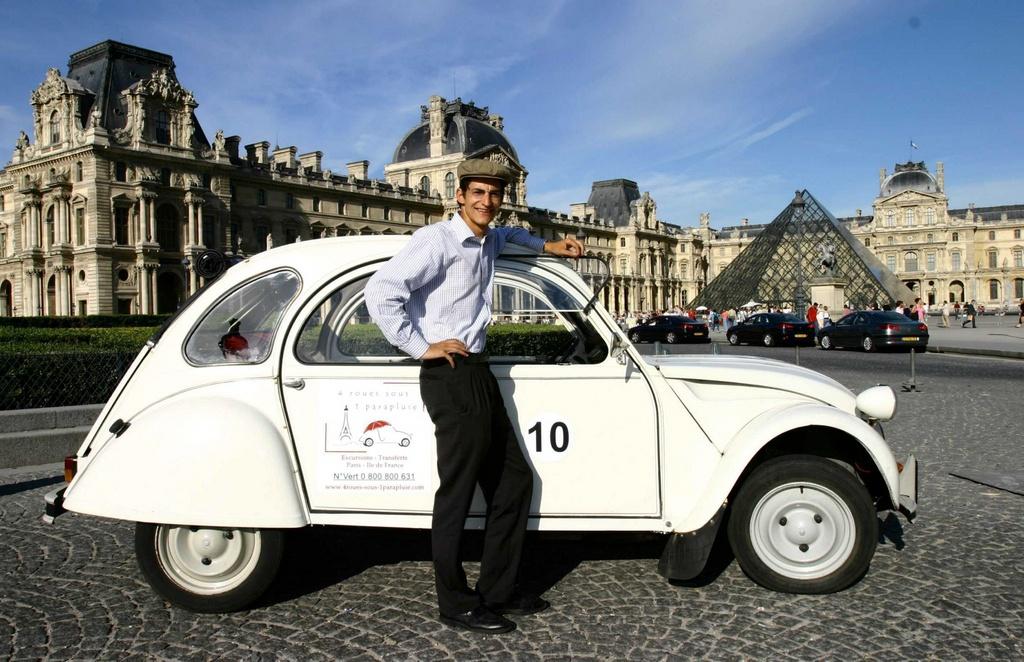 Un giovane davanti a una 2CV bianca davanti alla piramide di vetro, entrata del Louvre a Parigi.