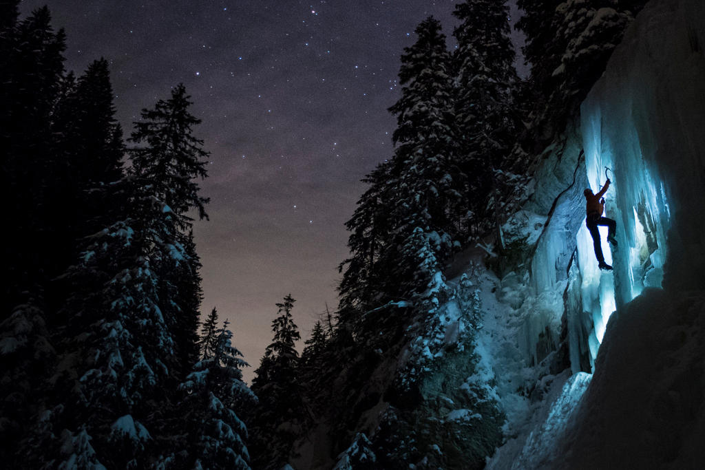 Uno scalatore in ascensione su una cascata di ghiaccio illuminata, in mezzo a una pineta, nella notte, sotto il cielo stellato.