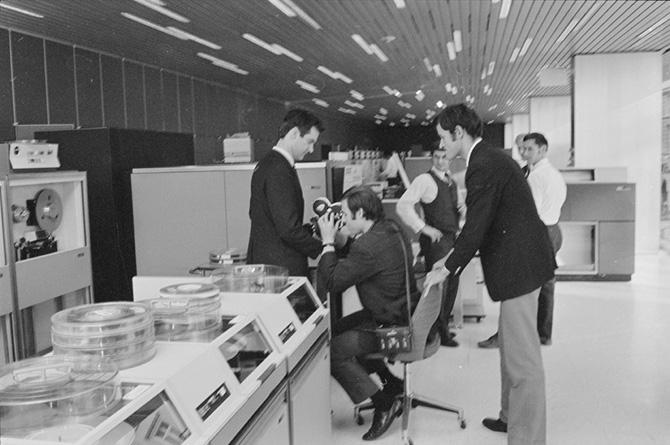 A group of men stand near a computer and one man is filming the computer.