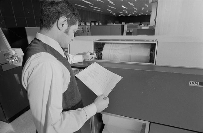 A man stands near a computer and reads from a print-out.