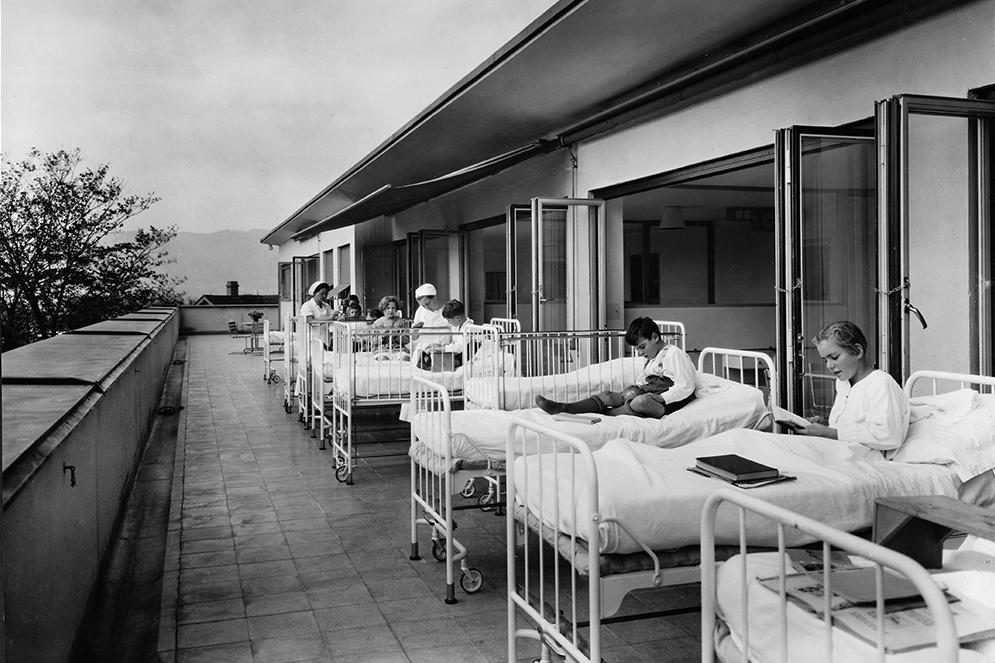 children sitting up in hospital beds, which are situated outside on the terrace of a building.