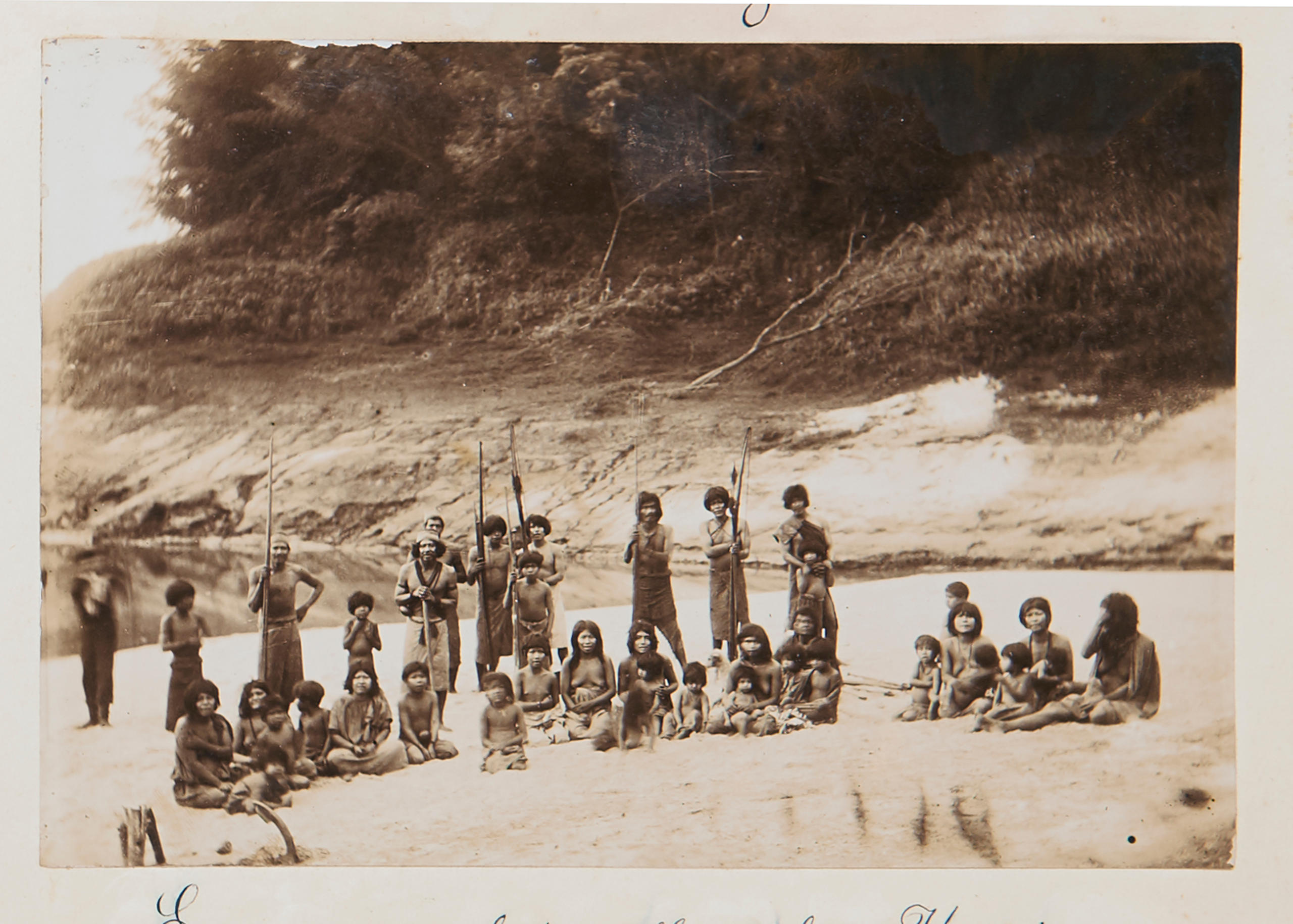 An old sepia photograph of a group of people standing (with spears) and sitting within the setting of a landscape.