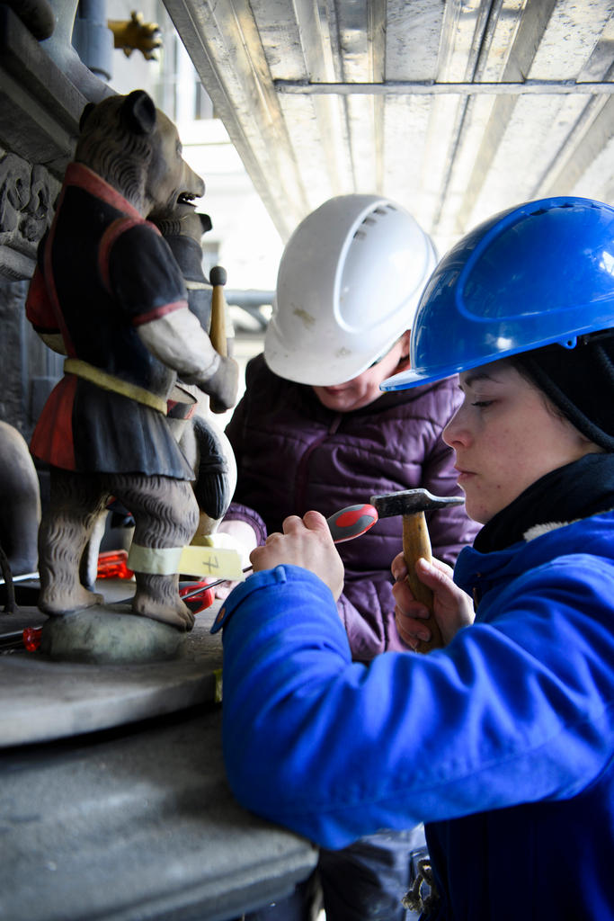 Two people wearing hard hats, one holding a small hammer while removing the figure of a bear.