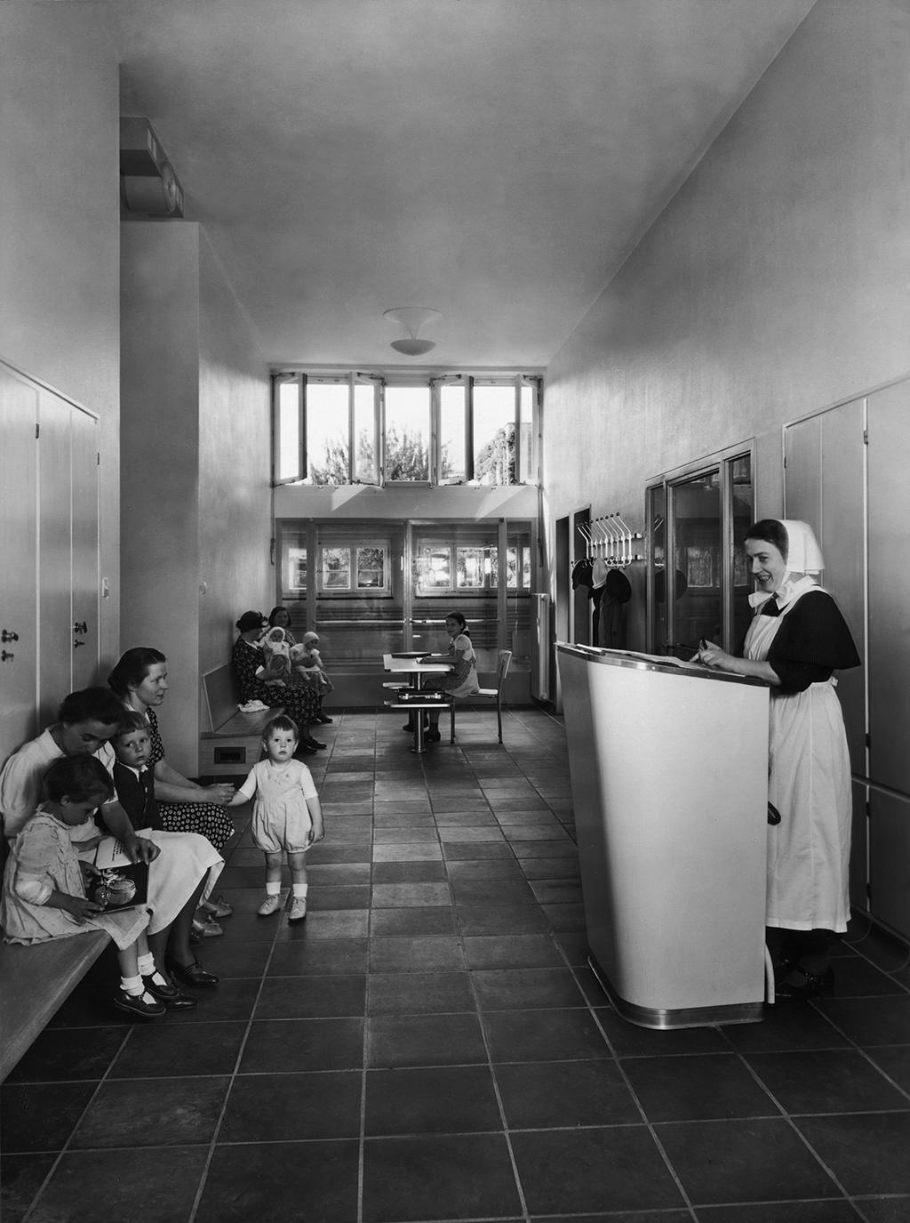 The entrance to a waiting area, children and women sitting to the left, to the right a woman dressed in a nurse uniform.