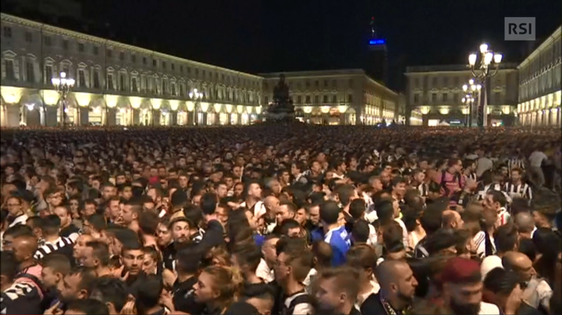 Folla in una piazza rivolta verso un maxischermo; parte delle persone si gira e si guarda alle spalle