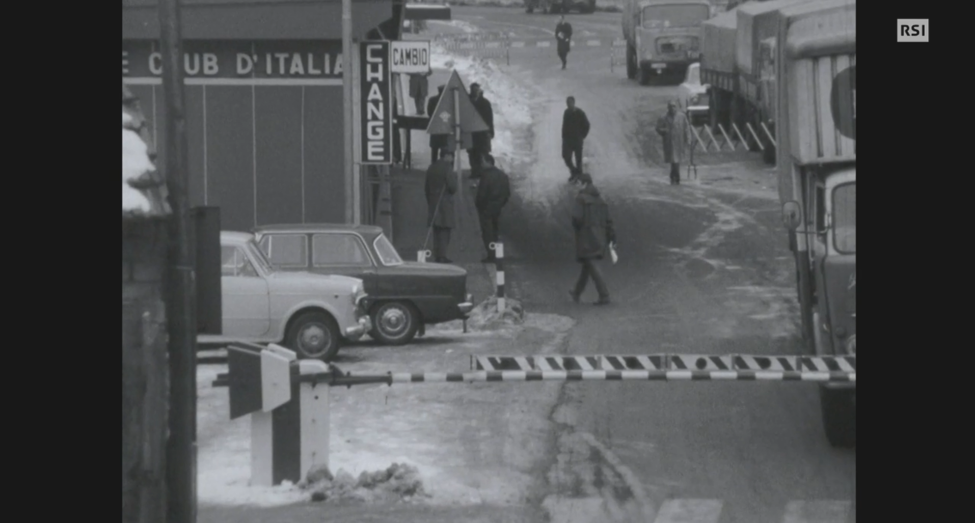 Una sbarra a righe (presumibilmente bianche e rosse) davanti a un posto di frontiera; persone attraversano a piedi