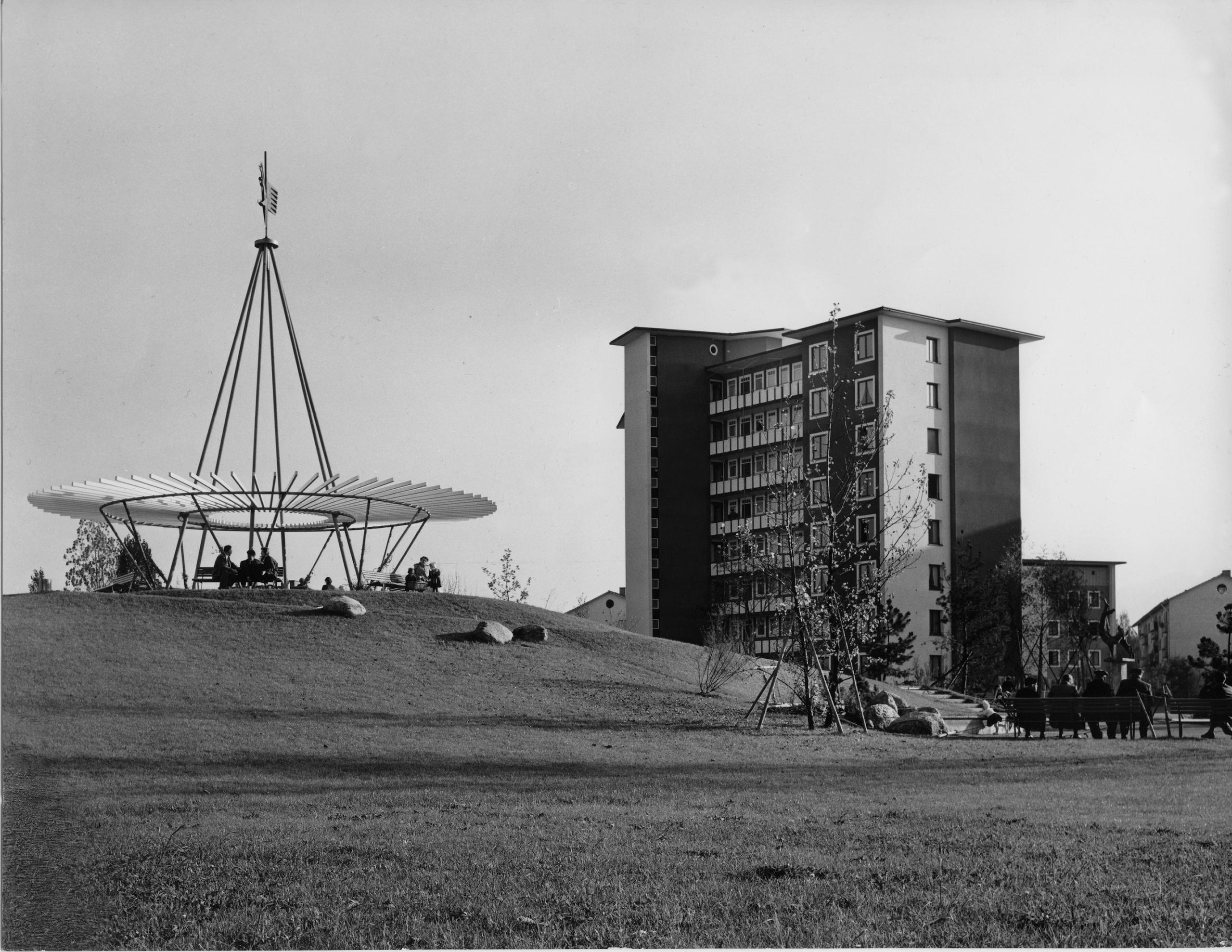 An apartment block to the right of a green field where are people sitting.