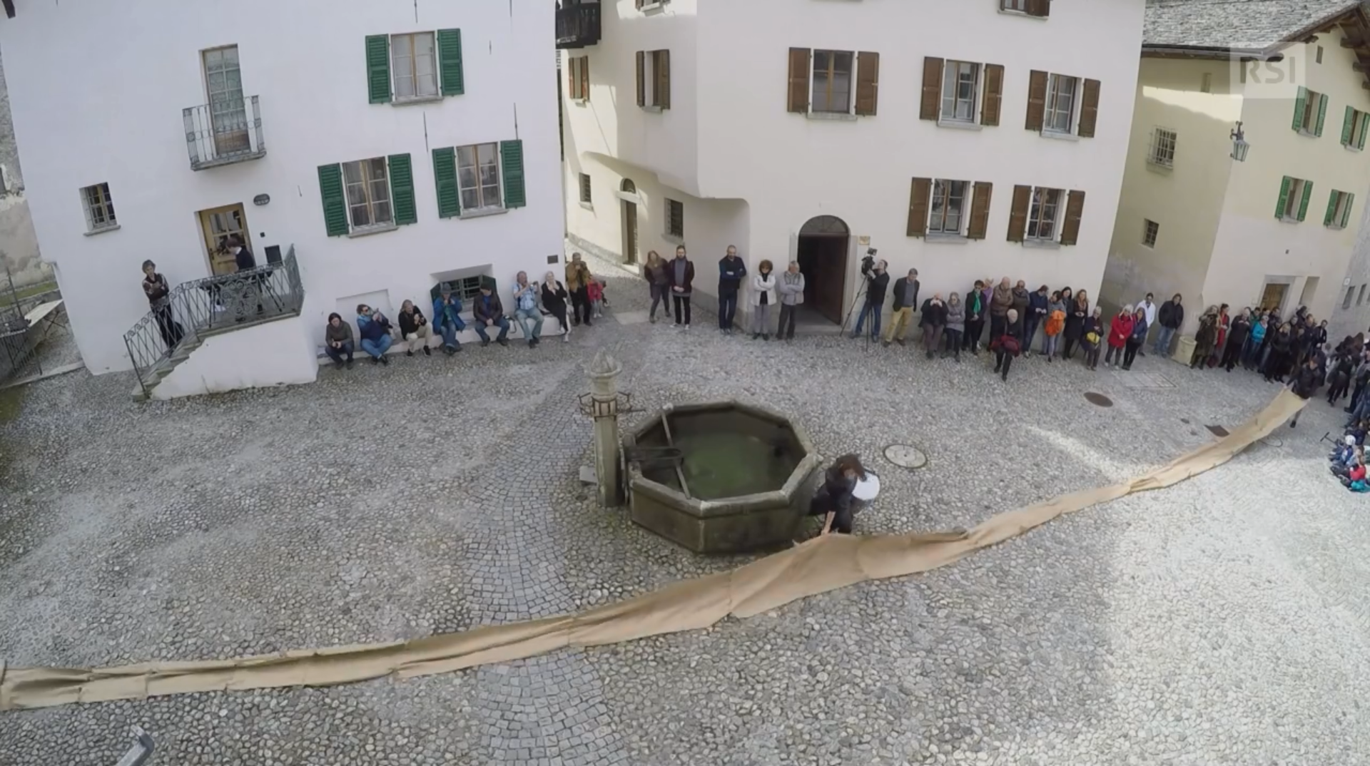 Una piazza di paese con pubblico ai margini e una figurante con tamburo al centro.