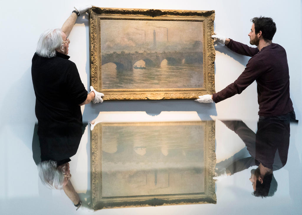 Two men wearing white gloves, hang a painting on the wall.