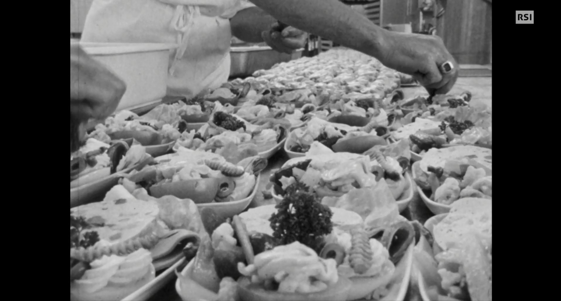 Tavolo di lavoro di una cucina industriale colmo di piatti di insalate fredde, una mano aggiunge ingredienti