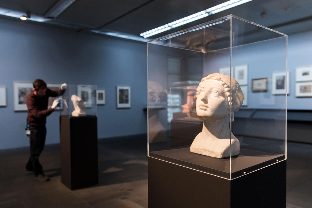 Foreground: a sculpture of a female head in a glass case within the setting of an art gallery or museum.