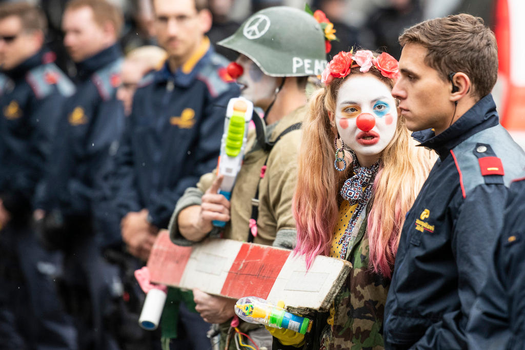 Manifestante vestita da clown parla a un impassibile poliziotto; sullo sfondo, sfocati, altri agenti