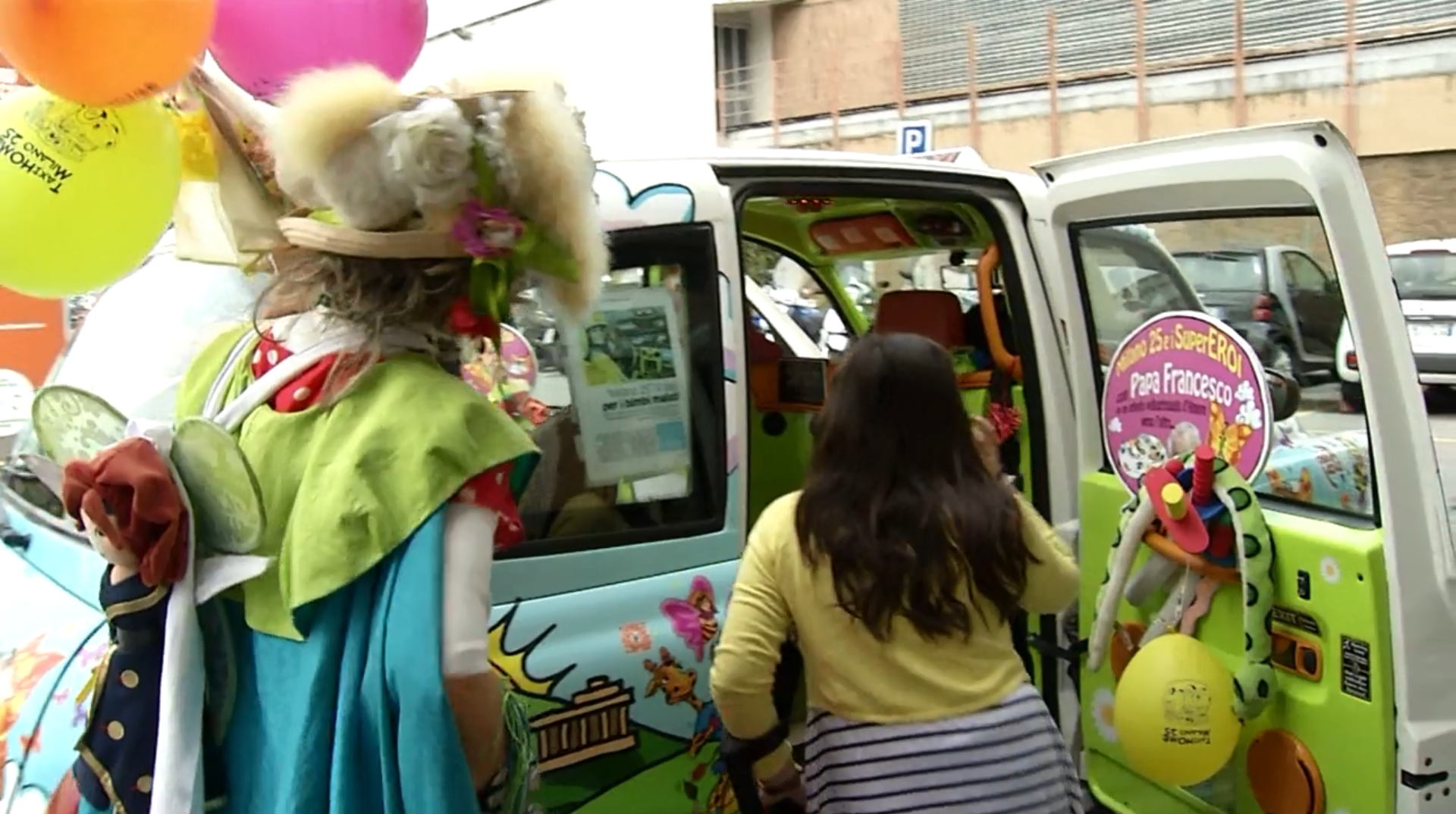Auto dipinta e decorata con peluche e palloncini, bambina sale a bordo. Di schiena, donna imparruccata con costume fiabesco