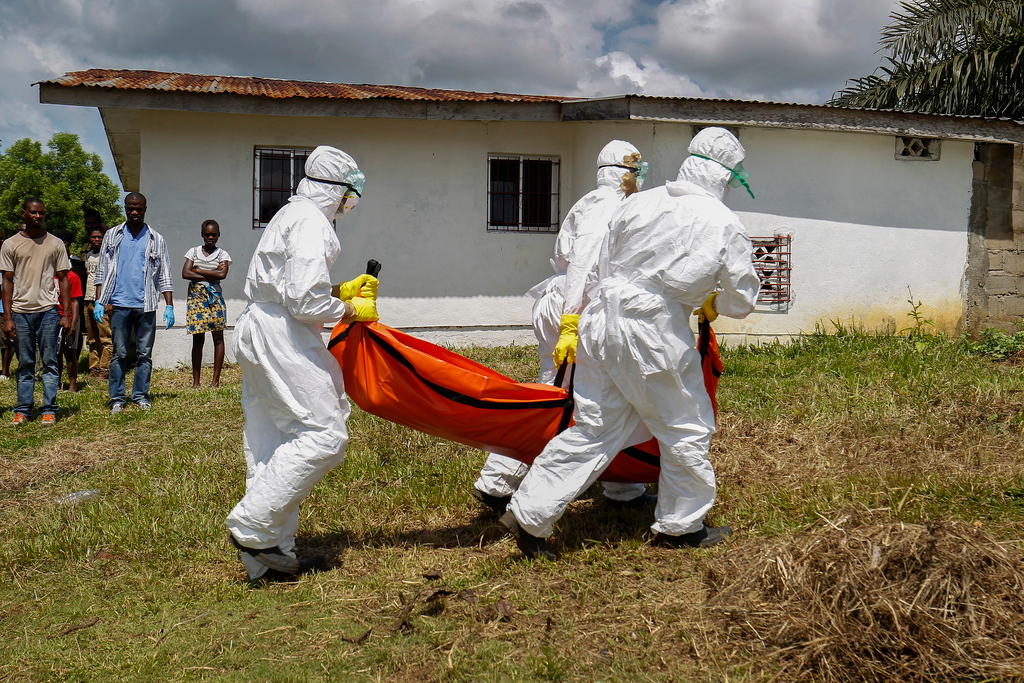 Tre soccorritori completamente coperti da tute bianche trasportano un cadavere avvolto in un manto arancione