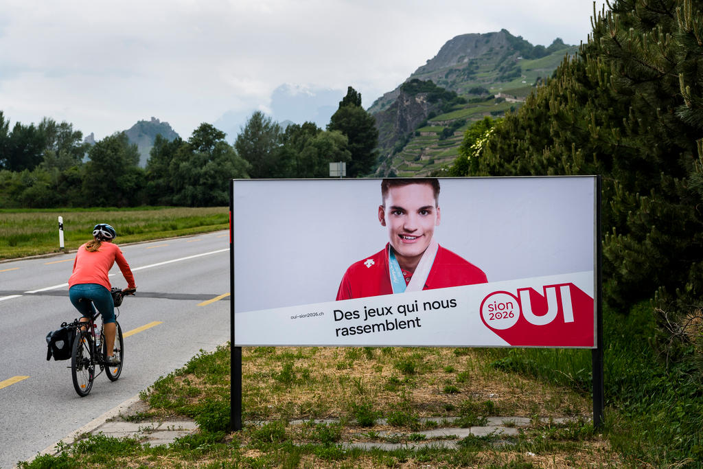 ciclista con a fianco un cartellone pubblicitario in favore di Sion 2026