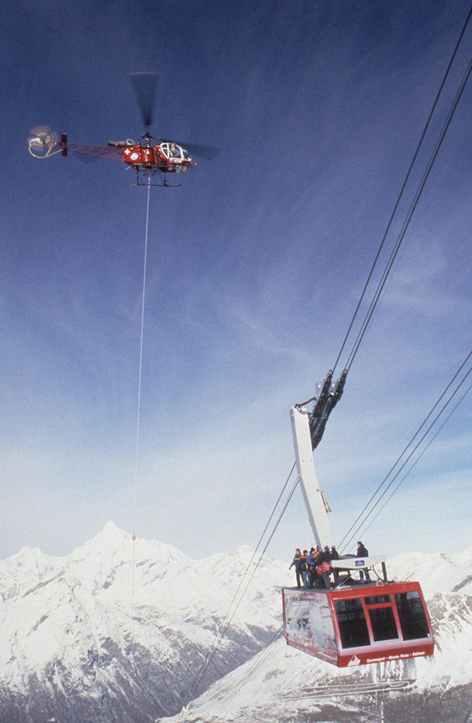 A helicopter flies above a cable car with people standing on its roof